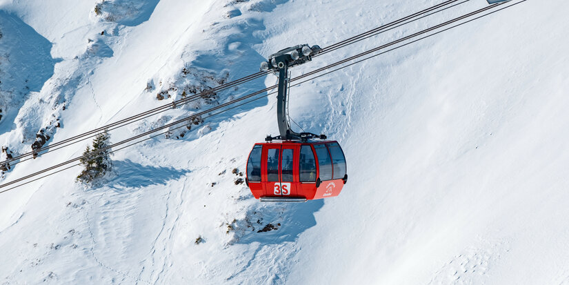 3S-Bahn, Skigebiet Kitzbühel, rote Gondel schwebt über verschneiter Landschaft mit Hügeln und Tannen.