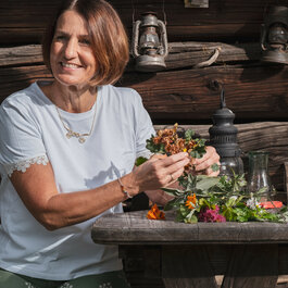 Eine Frau sitzt an einem Holztisch und arrangiert bunte Blumen, während sie freundlich in die Kamera lächelt.