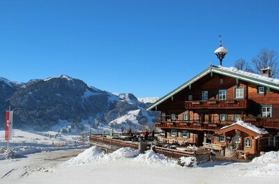 Rustikales Holzhaus mit Terrasse in einer verschneiten Berglandschaft unter blauem Himmel.