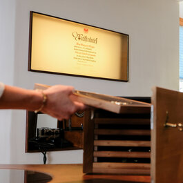 Eine Hand hält eine Schublade mit Schmuck, während im Hintergrund ein Meisterbrief an der Wand hängt.