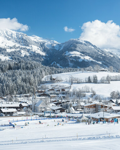 Reith, Skigebiet Kitzbühel, Dorf in verschneiter Landschaft mit Bergen bei sonnigem Wetter.