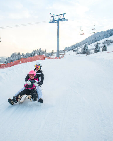 Skigebiet Kitzbühel, Erwachsener und Kind rodeln gemeinsam über verschneite Piste mit Tannen.