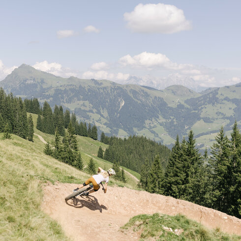 Ein Mountainbiker fährt mit Schwung auf dem Hahnenkamm Trail in Kitzbühel eine kurvenreiche Strecke durch die grüne Berglandschaft unter blauem Himmel.