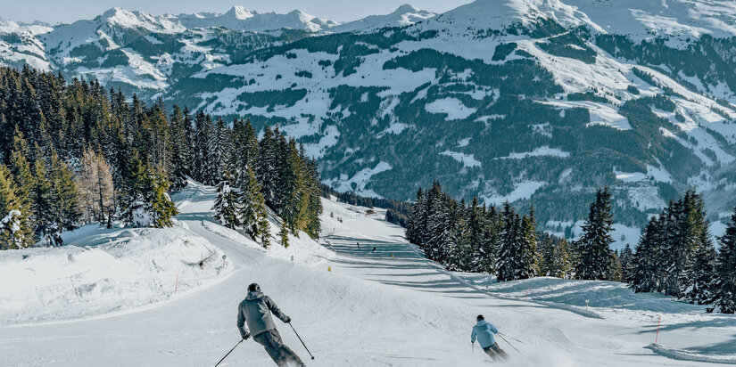 Jochberg, Skigebiet Kitzbühel – Zwei Skifahrer gleiten über eine schneebedeckte Piste, umgeben von bewaldeten Hängen und Bergblick.