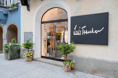 Eingangsbereich eines Luis Trenker Bekleidungsgeschäfts mit Rundbogentür und schwarzem Firmenschild.