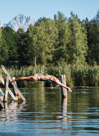 Der Schwarzsee in Kitzbühel und eine Frau, die einen Kopfsprung vom Steg aus hinein macht