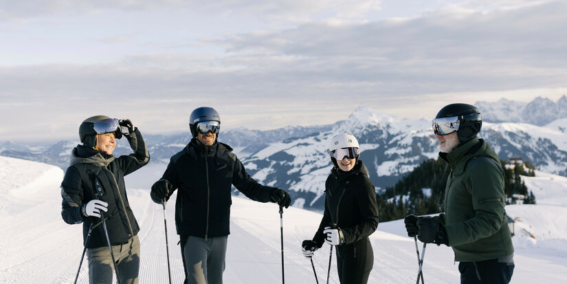 Skigebiet Kitzbühel – Vier Skifahrer stehen auf einer schneebedeckten Piste und blicken auf die Berge im Hintergrund.