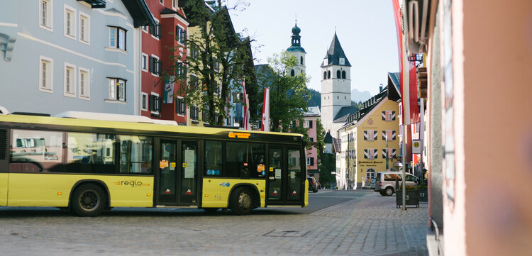 Ein gelber Bus fährt durch die malerische Innenstadt von Kitzbühel, umgeben von bunten Gebäuden und einem klaren Himmel.