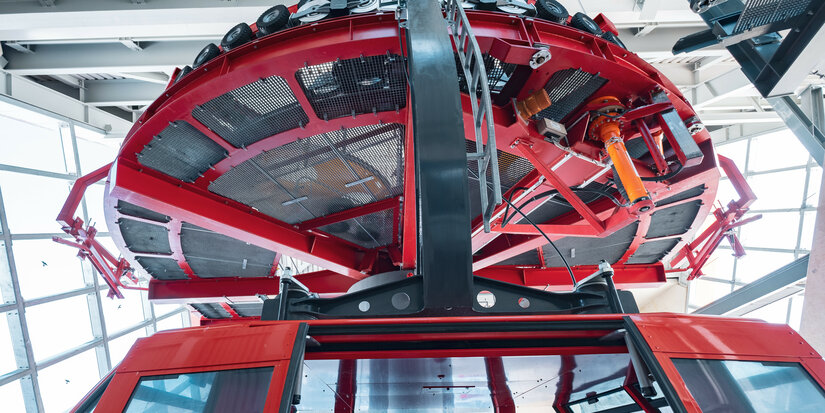 Die Unterseite einer roten Seilbahn mit einem großen, runden Dach und sichtbaren mechanischen Teilen im Innenraum.
