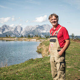 Ein lächelnder Mann in einem roten Shirt und Latzhose steht am Ufer eines klaren Sees, umgeben von majestätischen Bergen.