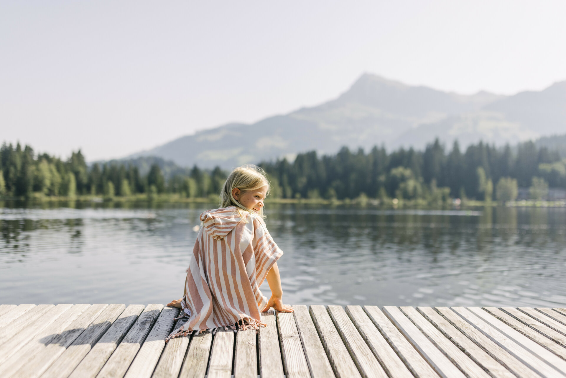 Ein kleines Mädchen sitzt am Ufer des Schwarzsees in Kitzbühel, umgeben von Bergen und Bäumen, und schaut lächelnd über das Wasser.