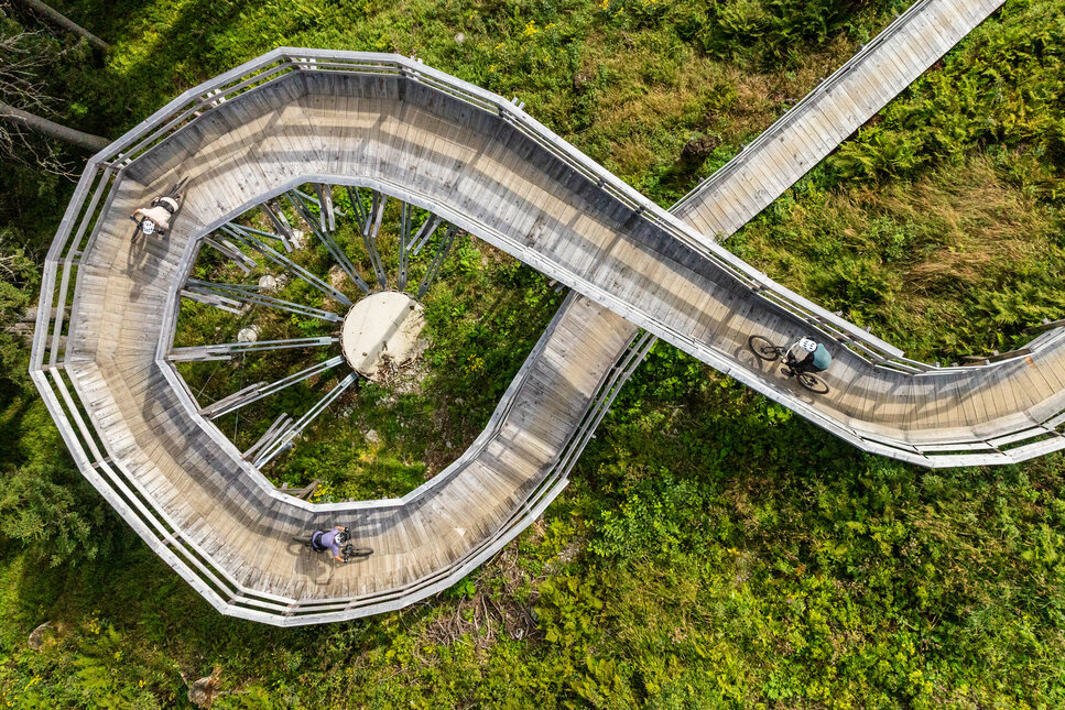 Eine geschwungene Holzbrücke am Sonnenrast Trail in Kitzbühel, umgeben von üppigem Grün, während Mountainbiker die Strecke nutzen.
