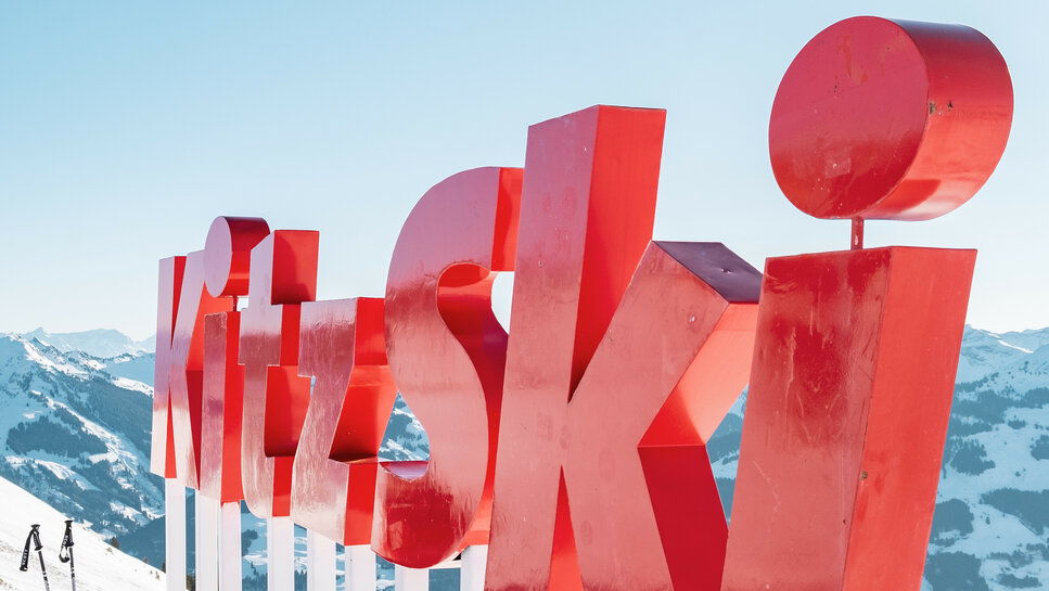 Ein leuchtend rotes Schild mit der Aufschrift 'KitzSki' steht auf schneebedecktem Boden, umgeben von majestätischen Bergen.