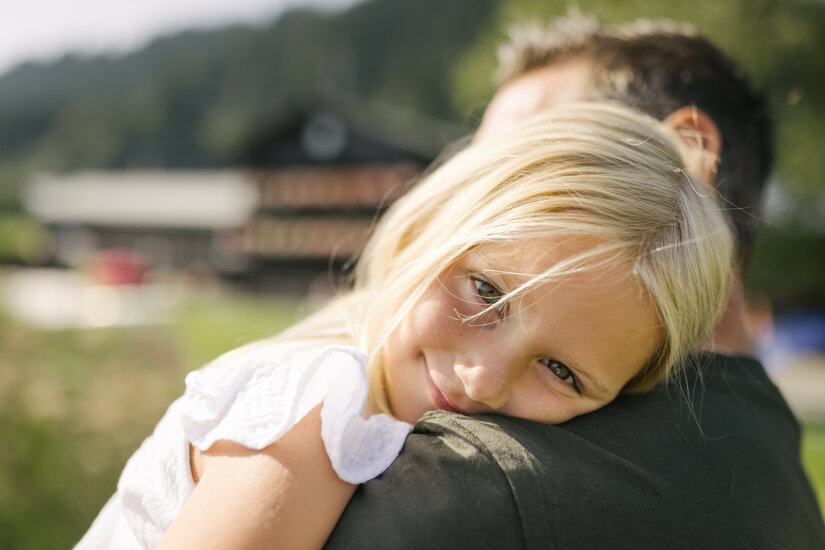 Ein kleines Mädchen mit blonden Haaren sitzt lächelnd auf dem Rücken eines Erwachsenen am Schwarzsee in Kitzbühel.