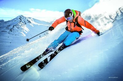 Skifahrer in orangefarbener Jacke und blauer Hose fährt eine steile, verschneite Piste hinunter.