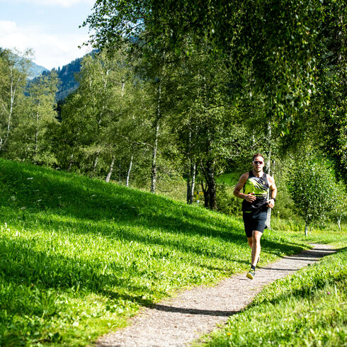 Ein sportlicher Mann joggt auf einem schmalen Weg durch eine grüne Landschaft mit Bäumen im Hintergrund.