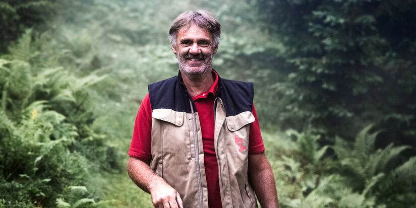 Ein lächelnder Mann steht in einem nebligen Wald, trägt eine Weste und hält einen Stock in der Hand.