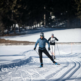 Beim Langlauf in Kitzbühel gleiten zwei Langläufer über verschneite Loipe, umgeben von dunklen Bäumen unter klarem Himmel.