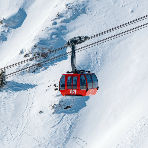 3S-Bahn, Skigebiet Kitzbühel, rote Gondelbahn über verschneiter Landschaft mit Tannen.