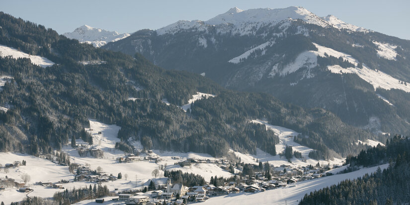 Winterlandschaft in Jochberg bei Kitzbühel mit Dorf, Bergen und Nadelbäumen.
