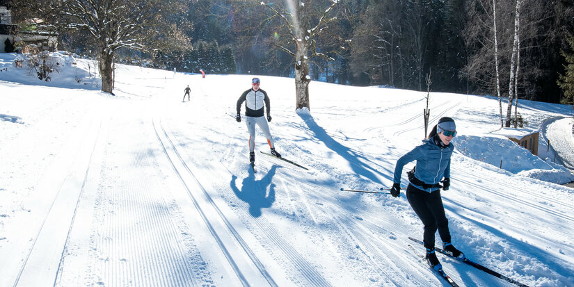 Beim Langlauf in Kitzbühel gleiten zwei Langläufer durch verschneite Landschaft im hellen Sonnenlicht mit Schattenwurf.