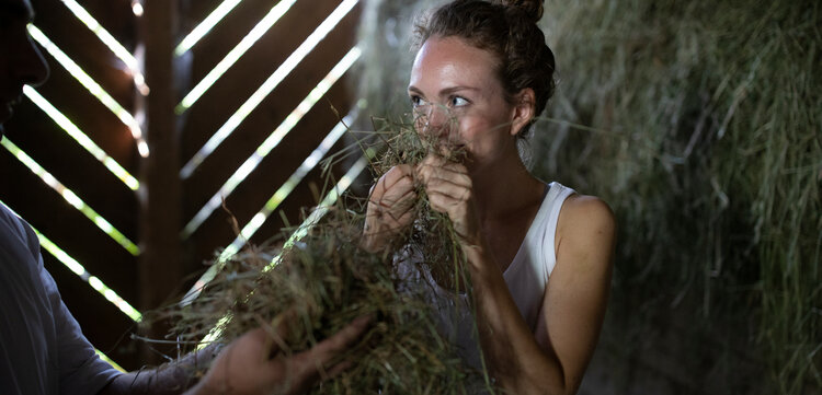 Eine junge Frau mit lockigem Haar hält Heu in den Händen und zeigt ein strahlendes Lächeln in einem Holzgebäude.
