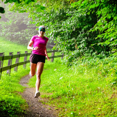 Eine Läuferin in einem pinken Shirt joggt auf einem schmalen Weg, umgeben von üppigem Grün und einem Holzgeländer.