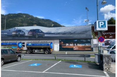 Behindertenparkplatz - Jochbergerstraße 27 Eine große Werbetafel zeigt zwei Autos vor einer Bergkulisse. Daneben steht ein Parkplatz mit einem Hinweis auf die Parkmöglichkeiten.