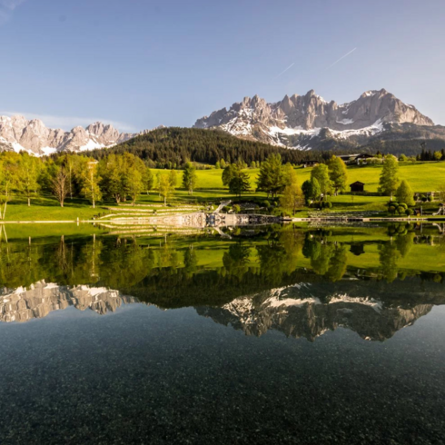 Ein ruhiger Badesee spiegelt die majestätischen Berge und den klaren Himmel wider, umgeben von üppigem Grün.