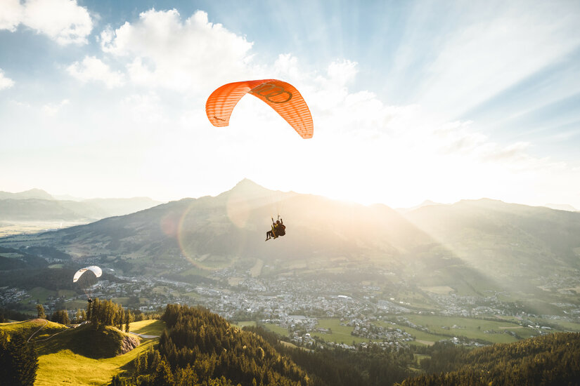 Ein Tandem-Paraglider fliegt über ein grünes Tal in Kitzbühel, umgeben von Bergen bei Sonnenschein.