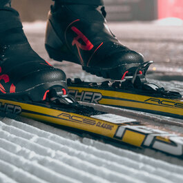 Beim Langlauf in Kitzbühel stehen schwarze Skischuhe mit roten Akzenten auf gelben Langlaufskiern in frisch präparierter Spur.