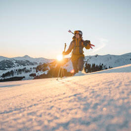 In der winterlichen Berglandschaft wandert eine Frau mit Skistöcken, während die Sonne den Himmel in warmen Farben erleuchtet.