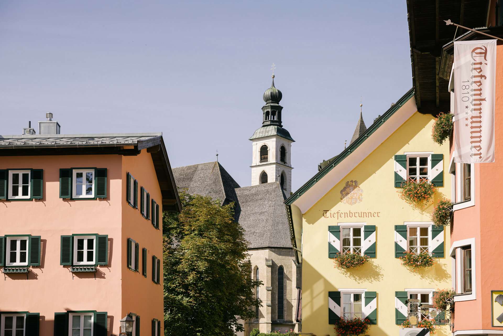 Bunte Häuser mit grünen Fensterläden und blühenden Balkonkästen, im Hintergrund der Kirchturm einer historischen Kirche.