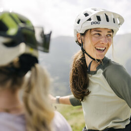 Eine lächelnde Frau mit Fahrradhelm genießt bei den 360 Flow Trails in Kitzbühel die Berglandschaft, während sie sich mit Freunden unterhält.