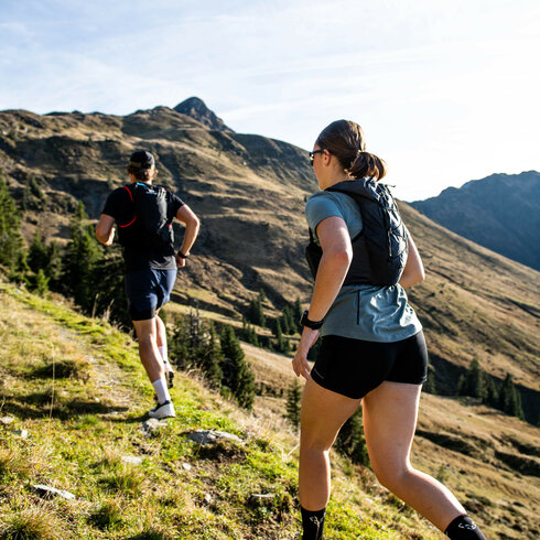 Zwei Trailrunner joggen energisch einen steilen Pfad in den Bergen, umgeben von grünen Wiesen und majestätischen Gipfeln.