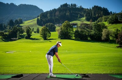 Ein Golfer steht auf einer Driving Range inmitten einer grünen Landschaft. Im Hintergrund sind sanfte Hügel und Bäume zu sehen.