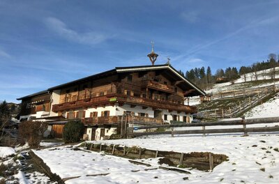 A traditional chalet-style house in a snow-covered landscape. The surrounding hills are green and the mountains are visible in the background.