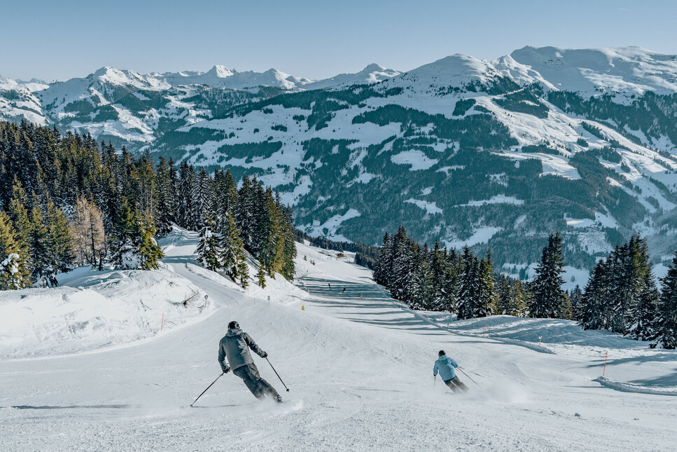 Jochberg, Skigebiet Kitzbühel, zwei Skifahrer auf verschneiter Piste mit Bergkulisse und Tannen.