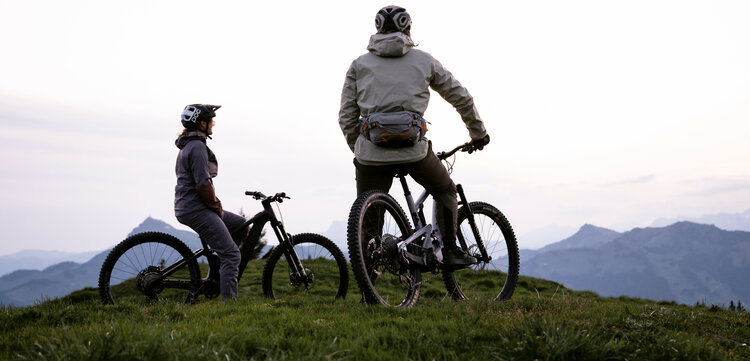 Zwei Mountainbiker stehen am Hahnenkamm in Kitzbühel und genießen bei Dämmerung den Blick auf die umliegenden Berge.
