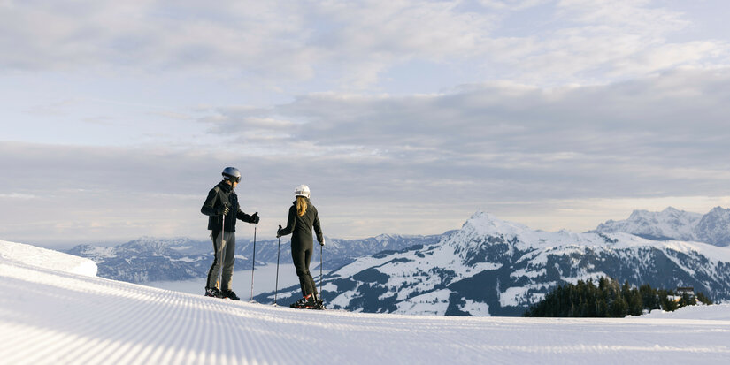 Skigebiet Kitzbühel – Zwei Skifahrer stehen auf einer schneebedeckten Piste und blicken über Berge und Tal.