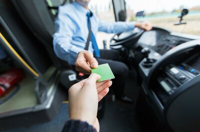 Ein Fahrgast reicht dem Busfahrer eine grüne Fahrkarte. Im Hintergrund ist das Innere eines Busses sichtbar.