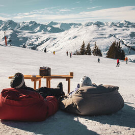 Skigebiet Kitzbühel, zwei Personen entspannen auf Sitzsäcken im Schnee mit Skifahrern im Hintergrund.