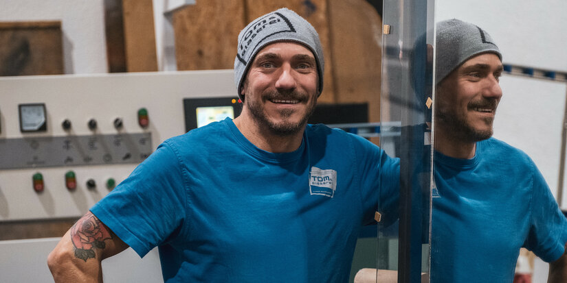 Der Mann mit einem grauen Beanie und einem freundlichen Lächeln posiert in einer Werkstatt mit Maschinen im Hintergrund.