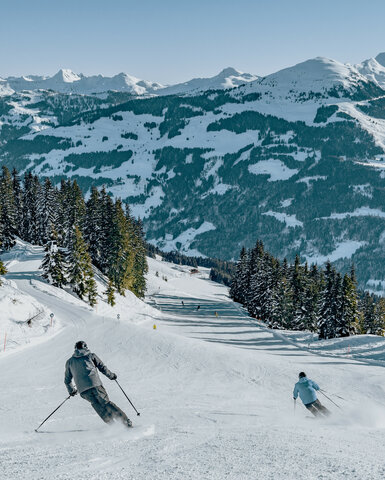 Jochberg, Skigebiet Kitzbühel, zwei Skifahrer auf verschneiter Piste mit Bergkulisse und Tannen.