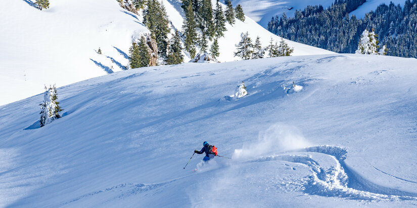 Im Skigebiet Kitzbühel gleitet ein Skifahrer durch frischen, unberührten Schnee in einer winterlichen Landschaft mit Bäumen im Hintergrund.