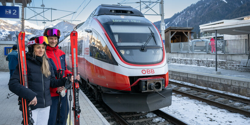 Bahnhof Hahnenkamm, Skigebiet Kitzbühel, Paar mit Skiern am Bahnsteig, moderner Zug im Hintergrund.