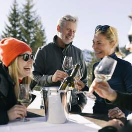 Skigebiet Kitzbühel, vier Freunde genießen Wein auf sonniger Terrasse vor verschneiten Bergen.