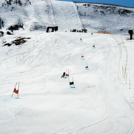 Skimovie-Strecke, Skigebiet Kitzbühel – Skifahrer fahren durch einen Riesentorlauf auf verschneiter Piste unter klarem, blauem Himmel.