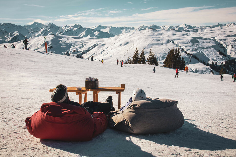 Skigebiet Kitzbühel – Zwei Personen in Sitzsäcken im Schnee mit Skifahrern im Hintergrund.