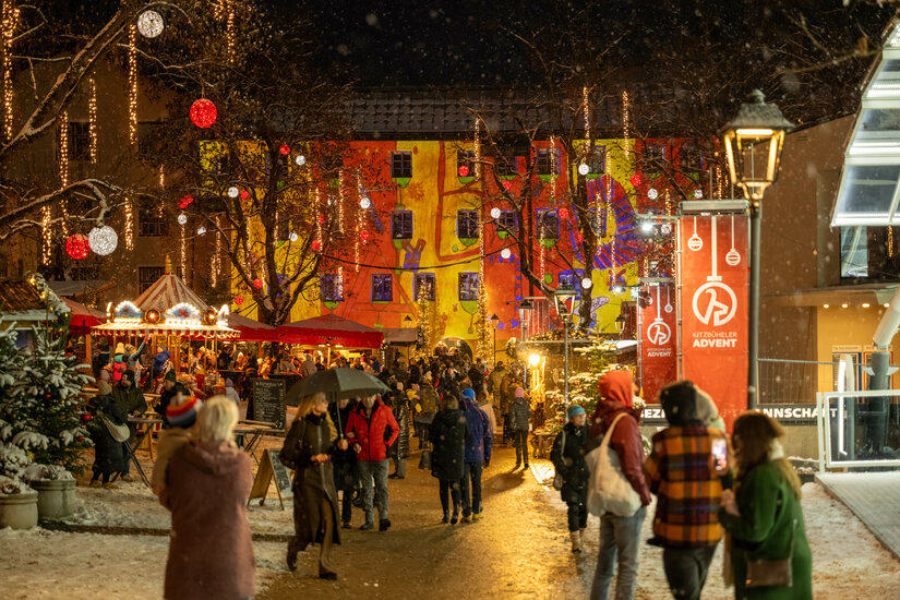 Eine festlich beleuchtete Straße in Kitzbühel, auf der Menschen durch den Weihnachtsmarkt schlendern, umgeben von bunten Lichtern und Schnee.